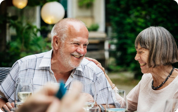 An older couple sit at an outdoor dining table, smiling and laughing with each other.