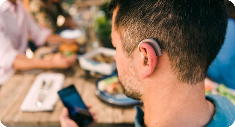 An image of a man looking at his cell phone. He is hearing a behind the ear hearing aid over his left ear. 