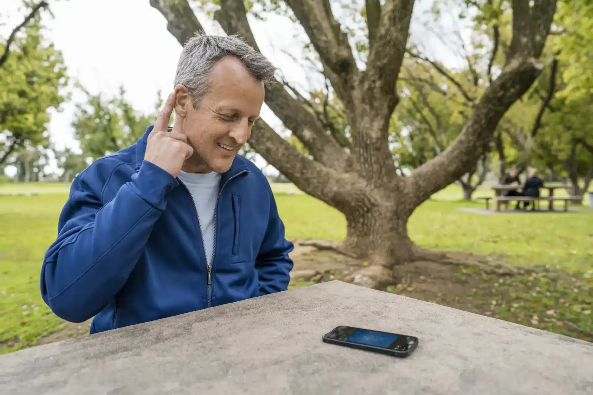 A man sitting at a park bench holds a finger up to his ear, tapping his hearing aid to take a phone call.